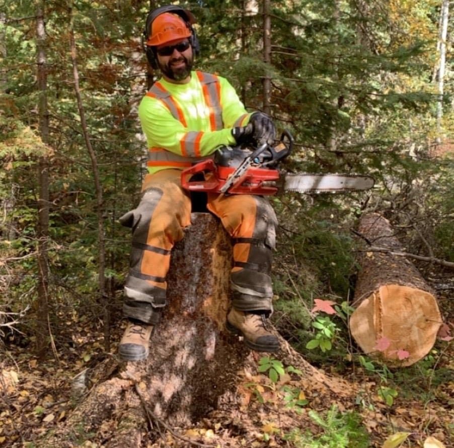 default-20 A bearded man in protective gear poses with a chainsaw atop a tree stump, next to a fallen tree.