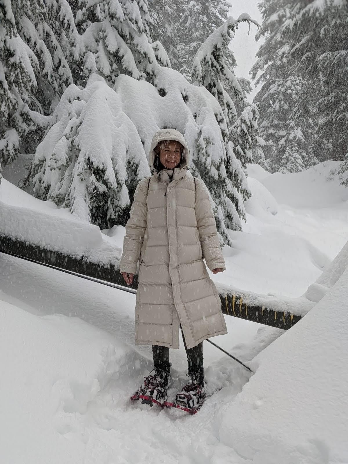 default-37 A woman in a white poofy jacket and snowshoes smiles as she poses on a snow-covered trail through tall pine trees.