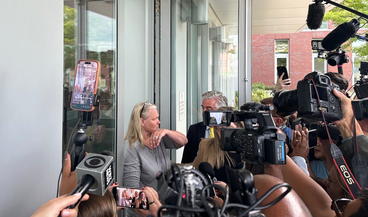 default-452 A woman with long, silver hair in a grey top exits a building surrounded by cameras and microphones.
